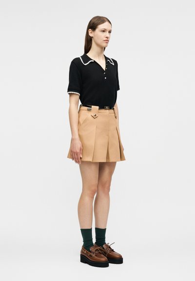 Γυναίκα που στέκεται φορώντας μαύρο τοπ με κοντά μανίκια και άσπρη διακοσμητική γραμμή, καμηλό πλισέ μίνι φούστα με ζώνη, σκουροπράσινες κάλτσες και καφέ παπούτσια με πλατφόρμα.