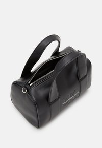 Sac à main en cuir noir Calvin Klein Jeans avec deux poignées et une fermeture éclair argentée partiellement ouverte montrant l'intérieur.