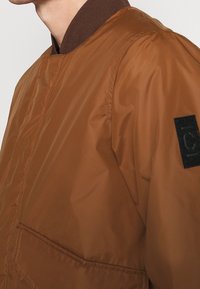 Brun bomberjacka med en slät, glänsande yta, med en brun ribbad krage och en svart patch på ärmen.