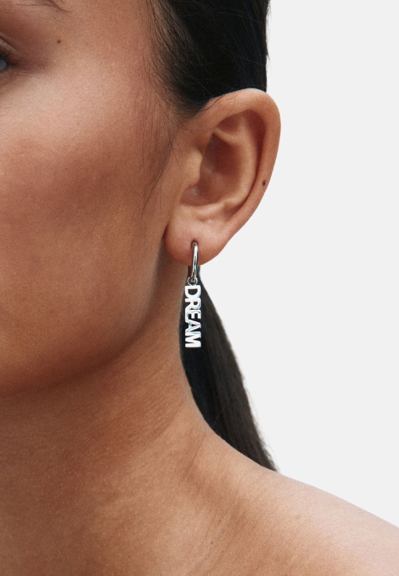 Sidabrinė lankelio formos auskaras su žodžiu „DREAM“ ryškiais didžiosiomis raidėmis, kabantis ant mažo užsegimo. Lygi paviršiaus struktūra ir poliruotas paviršius.