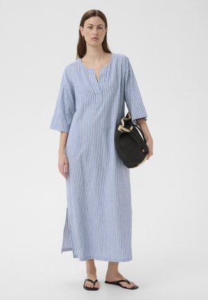 Femme portant une longue robe à rayures verticales bleues et blanches, tenant un sac noir tressé, et portant des tongs noires sur un fond uni.