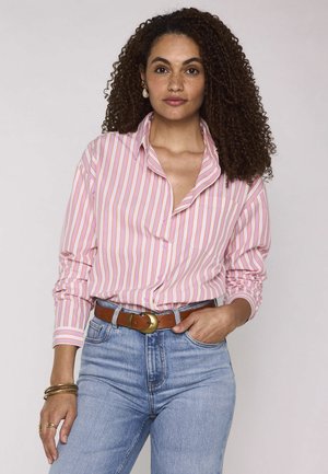 Donna con capelli ricci che indossa una camicia rosa e bianca a righe infilata in jeans blu chiaro con una cintura marrone, in piedi con una mano in tasca.