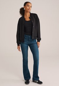 Črna bomber jakna s pinstripi in z gumbi na pritisek, v kombinaciji s temno modrimi širokimi džinsami in črnimi supergami. Pod njo je preprost črn top.