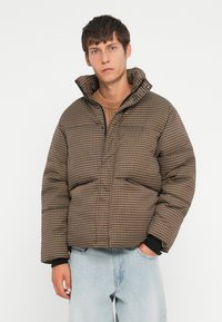 Bruine geruite pufferjack met hoge kraag en rits aan de voorkant, met twee voorzakken en elastische boorden, gecombineerd met lichtblauwe jeans.