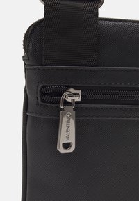 Detailní záběr na černý zip s texturou tašky se stříbrnou t�áhlicí s vyrytým nápisem "Valentino" a černým látkovým popruhem nad ním.