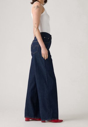 Personne portant un jean large bleu foncé, un haut blanc sans manches et des chaussures rouges, debout de profil avec des tatouages visibles sur le bras gauche.