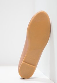 Helle, beige flache Schuhsohle mit einer strukturierten GummOberfläche, abgerundeter Form und geprägtem Branding. Glatte obere Kante geht in eine flache Basis über.