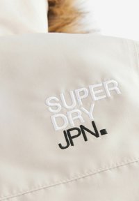 Tissu beige avec une texture lisse, ornée d'un logo brodé : "SUPER DRY" en blanc et "JPN." en noir. Détails de couture visibles.