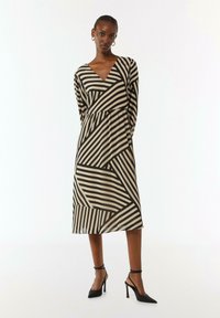 Langes V-Ausschnitt Kleid mit einem geometrischen Muster aus schwarzen und cremefarbenen Streifen, langen Ärmeln und einer taillierten Taille, kombiniert mit schwarzen High-Heel-Schuhen.