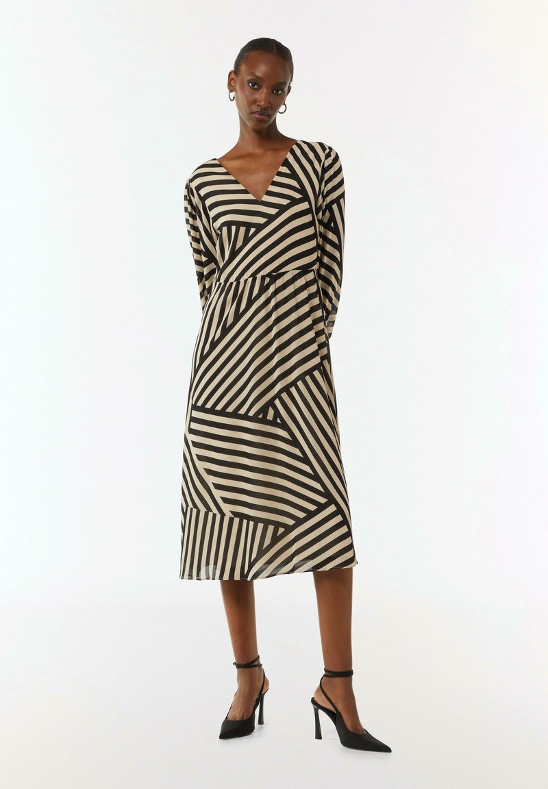 Langes V-Ausschnitt Kleid mit einem geometrischen Muster aus schwarzen und cremefarbenen Streifen, langen Ärmeln und einer taillierten Taille, kombiniert mit schwarzen High-Heel-Schuhen.