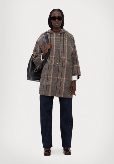 Casaco oversized de padrão xadrez castanho com capuz, apresentando fechos de botão e mangas até ao cotovelo, combinado com jeans escuros e sapatos brilhantes.