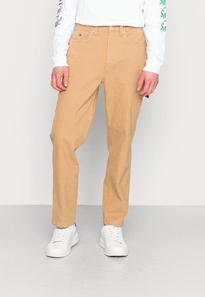 Person trägt beige gerade geschnittene Hose, weißes langärmeliges Hemd und weiße Turnschuhe, steht vor einem einfachen hellen Hintergrund.