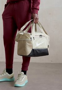 Une personne en pantalon de survêtement bordeaux et en baskets claires tient un sac de sport Nike beige et noir par les poignées contre un fond uni.