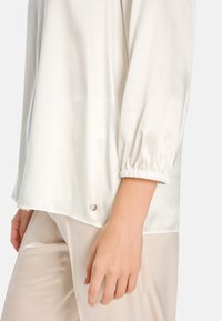 White satin blouse with long, gathered sleeves; smooth texture; features a small metallic logo near the hem. Lightweight and flowy design.
