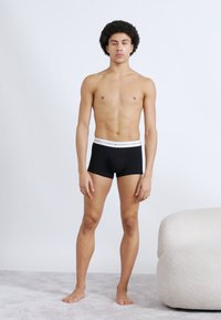 Μαύρα αντρικά εσώρουχα boxer με λευκή ελαστική μέση. Το μοντέλο στέκεται με καλή στάση μπροστά σε απλό φόντο, χωρίς επιπλέον αξεσουάρ.