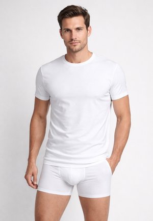 Uomo che indossa una maglietta aderente bianca a maniche corte e boxer bianchi, in piedi contro uno sfondo chiaro con una mano in tasca.