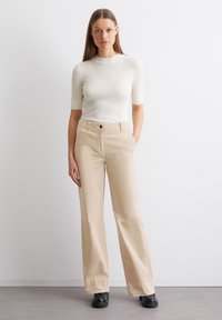 Pantalons larges beiges avec un devant plat, un bouton noir unique et des poches latérales, associés à un haut en tricot blanc ajusté à manches courtes.