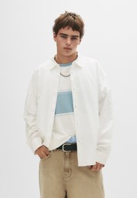 Weißes Hemd über einem blau-weiß gestreiften T-Shirt, kombiniert mit beigen Denim-Hosen und einem schwarzen Gürtel. Kurze, strukturierte Haare.