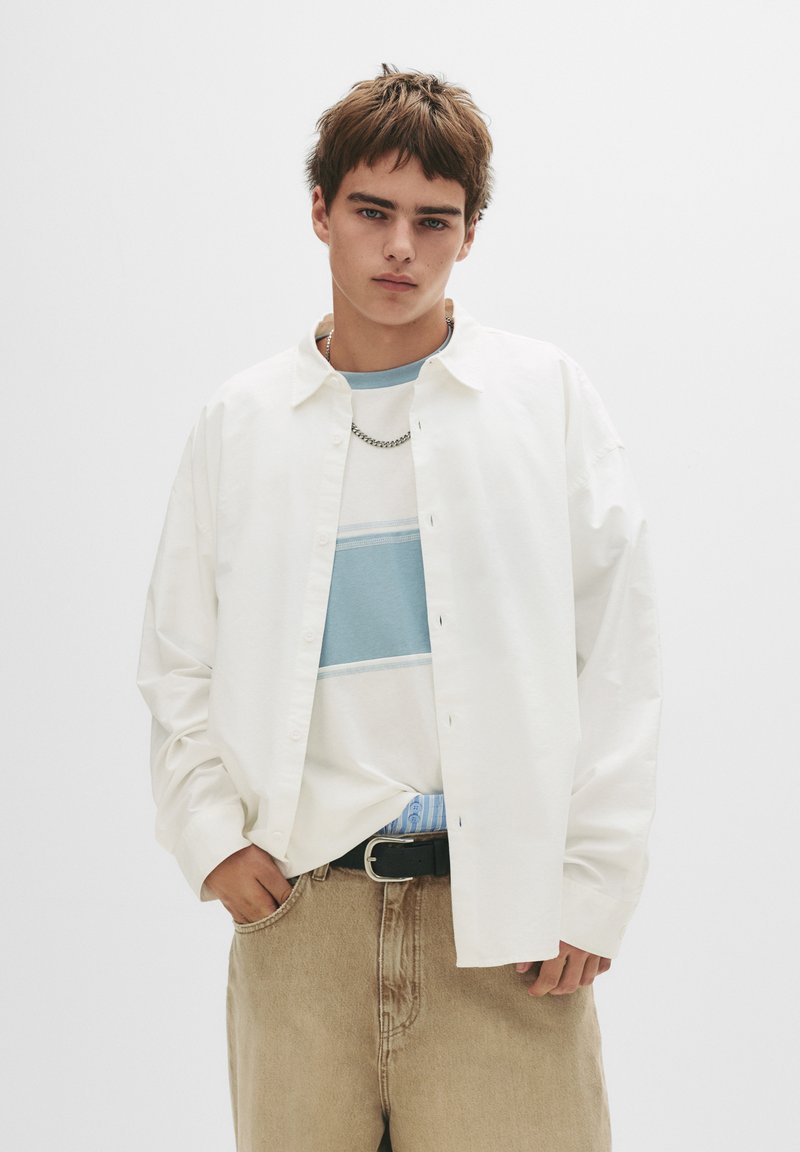 Weißes Hemd über einem blau-weiß gestreiften T-Shirt, kombiniert mit beigen Denim-Hosen und einem schwarzen Gürtel. Kurze, strukturierte Haare.