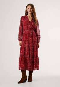 Vestido longo vermelho com padrão floral, mangas franzidas nos punhos, saia de camadas, feito de tecido leve, combinado com botas castanhas até ao joelho.