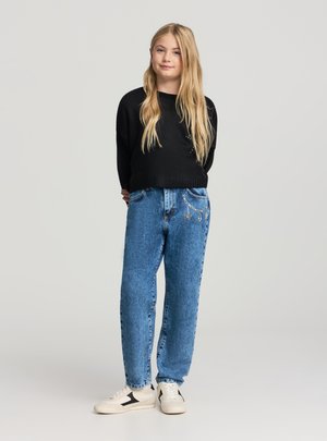 Ragazza giovane con lunghi capelli biondi, che indossa un maglione nero, jeans blu decorati con catene e sneakers bianche, in piedi con le mani dietro la schiena.