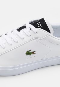 Fehér bőr sportcipő, alacsony felsőrésszel, zöld krokodil logóval, fehér fűzőkkel és fekete kiemeléssel a nyelvén. Texturált talp.