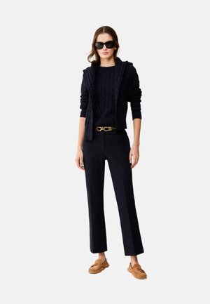 Vrouw draagt een zwarte gebreide trui, zwarte broek met gouden riem, bruine mocassins en zwarte zonnebril, staand tegen een witte achtergrond.