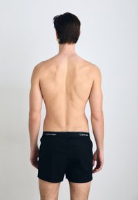 Črne bokserice z mehkimi pasom, ki nosijo logo "Calvin Klein". Material se zdi gladek; šortki so krojeno, sedenje nad stegnom.