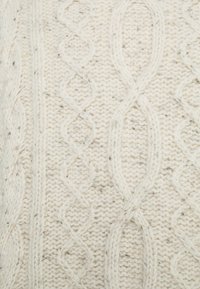Krämfärgad stickad tyg med en strukturerad kabelmönster och subtila inslag. Designen inkluderar snurrande linjer och öppna utrymmen.