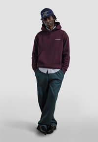 Felpa con cappuccio color marrone scuro con tasca frontale e logo bianco, indossata sopra una camicia a righe blu. Pantaloni verde scuro e scarpe nere completano l'outfit.