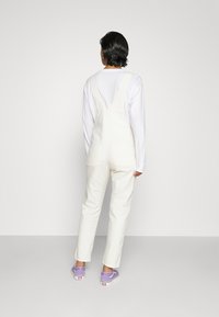 Salopette in denim bianco con spalline regolabili, dotata di tasche laterali, abbinata a una camicia bianca a maniche lunghe e scarpe slip-on viola.