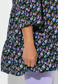 Sort blomsterkjole med livlige lilla og blå blomster, en gathered nederdel og flagrende ærmer. Hånd hvilende nær kanten, kombineret med lavendel leggings.