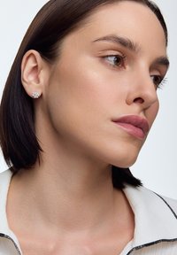 Okrągłe srebrne kolczyki z kwiatowym wzorem, ozdobione wieloma małymi kamieniami szlachetnymi. Teksturowana skóra na gładkim, neutralnym tle.