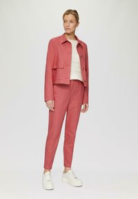 Roze cropped jack met knoopsluiting en borstzak, gecombineerd met bijpassende taps toelopende broek. Een witte gebreide trui en sneakers maken de look af.