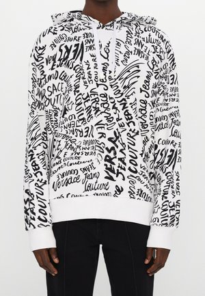 Personne portant un sweat à capuche blanc couvert de mots manuscrits noirs incluant Versace Jeans Couture, associé à un pantalon noir.