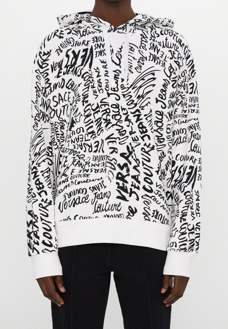 Personne portant un sweat à capuche blanc couvert de mots manuscrits noirs incluant Versace Jeans Couture, associé à un pantalon noir.