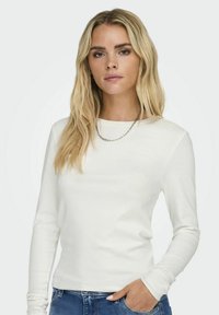 Chemise blanche à manches longues en tissu doux, avec un col rond et une coupe ajustée. Portée avec un jean en denim, mettant en avant un look décontracté.