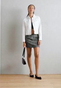 Chaqueta de mezclilla blanca, top corto negro, falda mini de patrón en blanco y negro, zapatos planos negros y bolso pequeño, de pie sobre un suelo de madera, contra una pared gris.
