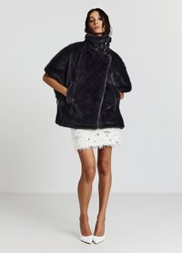 Manteau en fausse fourrure noir avec un col haut, des manches courtes et des poches latérales. Porté par-dessus une mini jupe blanche texturée, complété par des talons blancs.