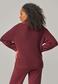 Frau mit lockigem Haar, die einen bordeauxroten, gerippten Rollkragenpullover und passende Hosen trägt, posiert mit den Händen in der Hüfte und blickt nach links.