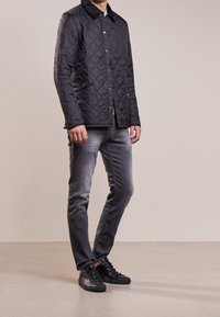 Veste noire matelassée avec un col en velours côtelé, des boutons de fermeture et deux poches latérales, associée à un jean gris et des baskets noires.