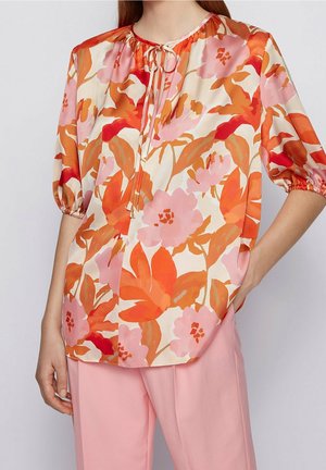 Femme portant un chemisier ample à fleurs orange et roses et un pantalon rose, une main dans la poche, sur un fond uni.