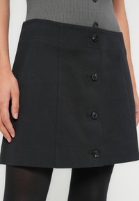 Μαύρη φούστα Α γραμμής με κουμπιά, με υφή, διαθέτει πέντε μαύρα κουμπιά μπροστά και λείο ζωνάκι στη μέση.