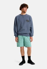 Felpa grigia con logo frontale, abbinata a pantaloni corti verde menta, calze nere e scarpe slip-on nere. Tessuto morbido con vestibilità rilassata.