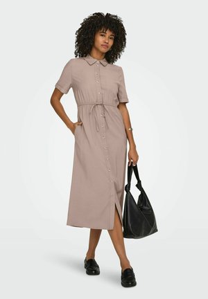 Görög hajú nő bézs színű, gombos midi ruhában, derekán kötővel, fekete loaferekkel, ezüst karkötővel, kezében fekete táskával.