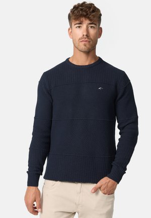 Ung mand med kort krøllet hår iført en mørk marineblå tekstureret striktrøje og lyse beige bukser, stående med den ene hånd i lommen.