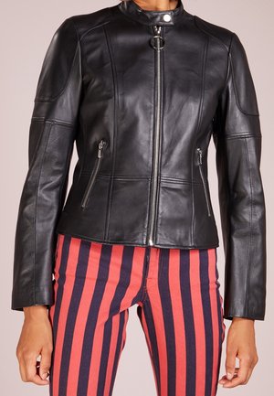 Veste en cuir noire zippée avec col montant et poches zippées, portée avec un pantalon à rayures verticales rouges et bleu marine.