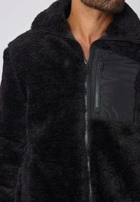 Homme portant une veste en polaire noire avec un col haut et une poche poitrine zippée en tissu doux.