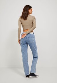 Beige hosszú ujjú crop top, kék, magas derékú farmernadrág átlátszó zsebekkel, és fekete sportcipők. Az alak hátulról látható.