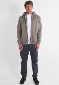 Sweat à capuche zippé gris avec cordons de serrage, doublure intérieure et design brodé sur la poitrine, associé à un pantalon gris foncé et des baskets noires.